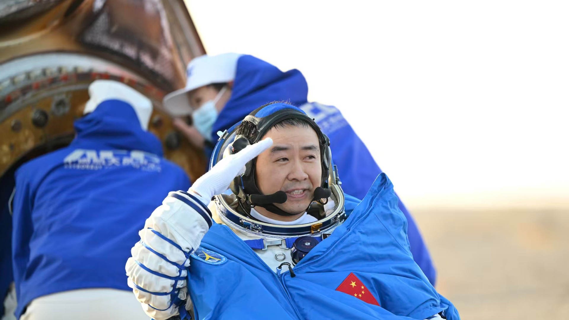 a scorched cone-shaped capsule lies on a desert floor surrounded by people in thick blue coats