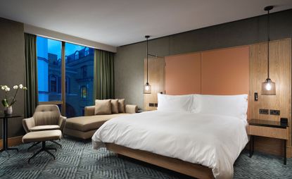 A room in the Hilton London Bankside UK. Spacious room in gray, beige, and orange tones. Large bed with a tall headboard in a combination of wood and leather. To the left, there is a large window, with a couch and a chair in front of it.