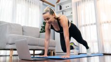 A woman performs a low impact burpee at home. She is on an exercise mat, her arms straight and hands on the mat, with her left leg stepped back straight behind her. Her right knee is bent as she draws her right foot forward. Behind her we see a couch and bookshelves. 