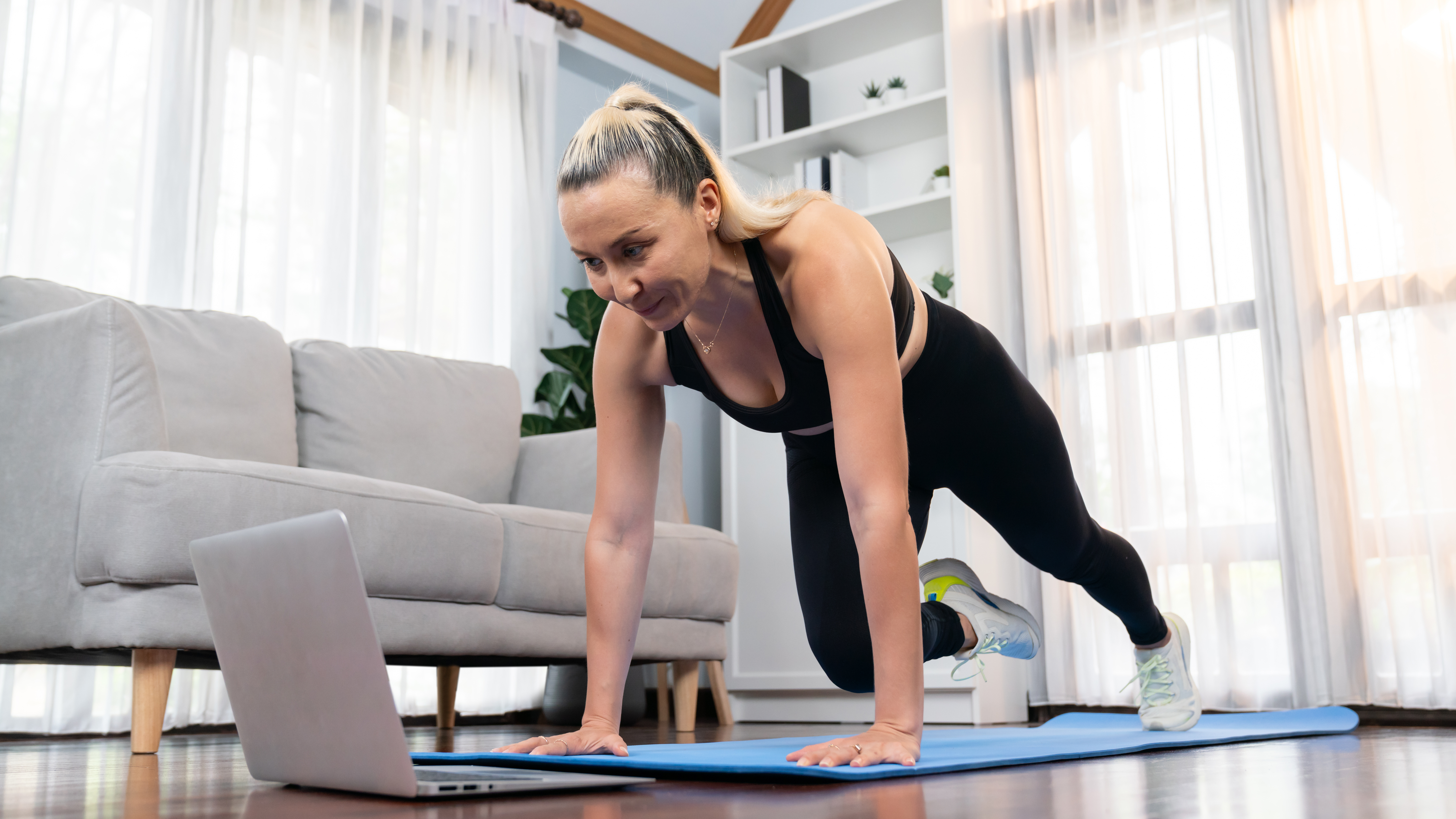 A woman performs a low impact burpee at home. She is on an exercise mat, her arms straight and hands on the mat, with her left leg stepped back straight behind her. Her right knee is bent as she draws her right foot forward. Behind her we see a couch and bookshelves. 
