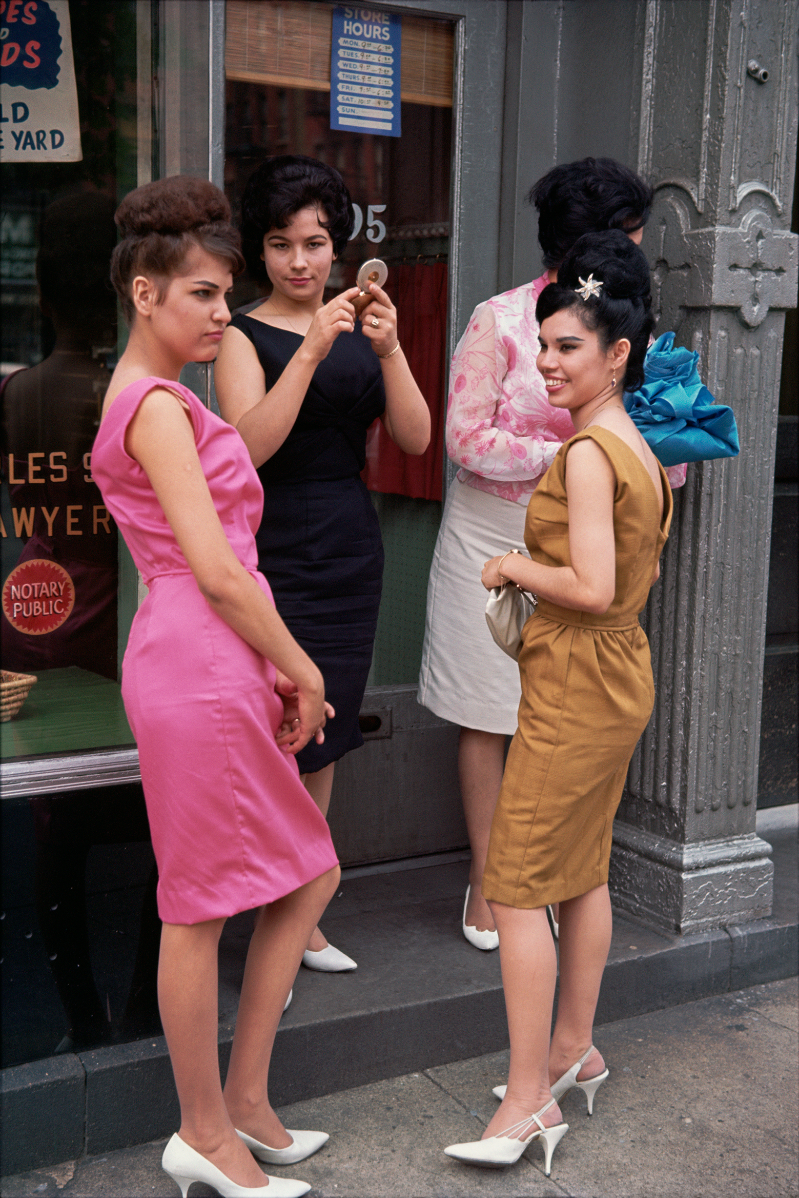 A group of four women in colorful 1960s dresses stand on a sidewalk, engaged in conversation outside a storefront