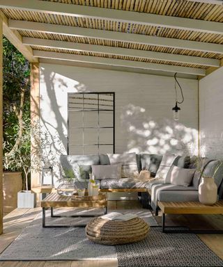 Outdoor porch area with bamboo ceiling, white painted brick walls, gray corner sofa, two outdoor rugs, woven, textured ottoman
