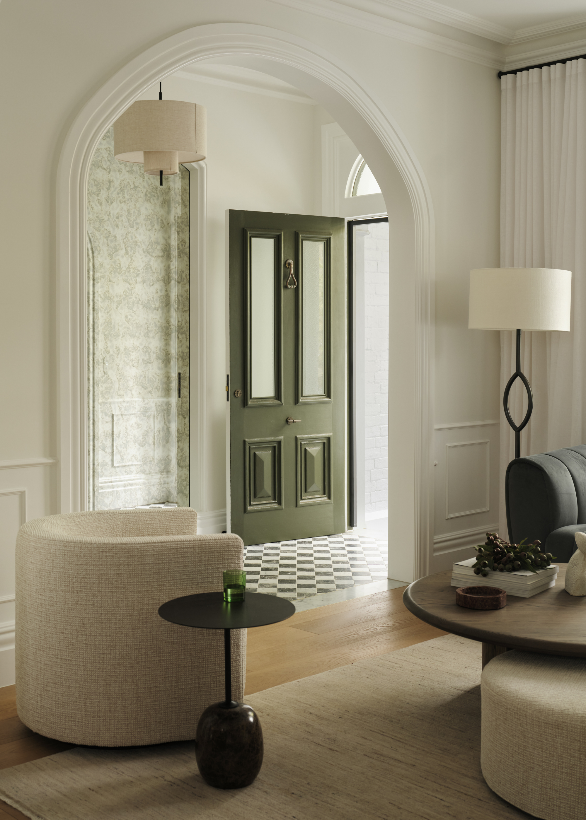 A living room with an arched doorway with millwork, a checkerboard green stone floor, a black floor lamp with a white shade, a curved sofa chair, a black side table with a green glass, a coffee table with grapes, and a beige rug on the floor