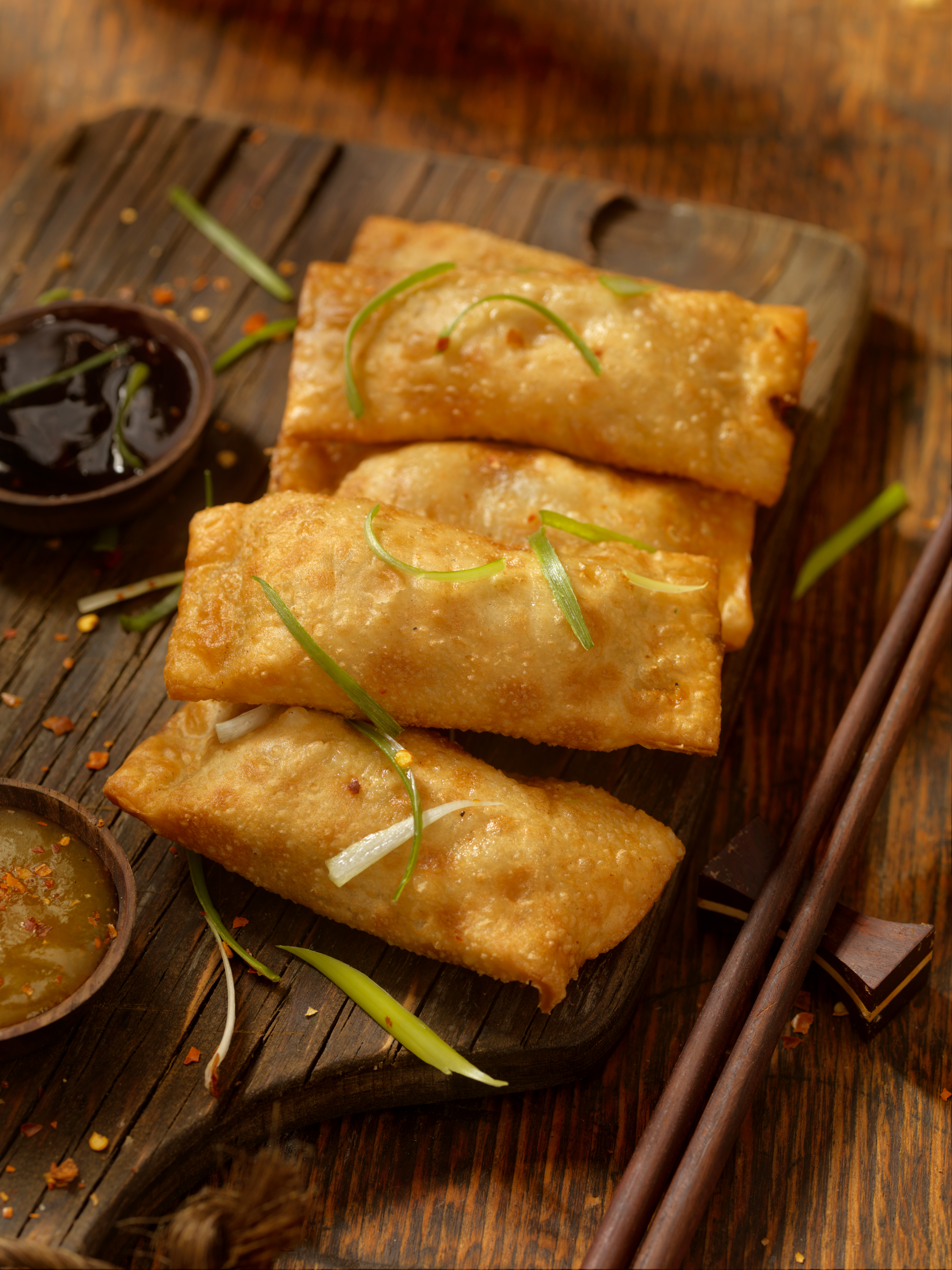 A platter of fried egg rolls, sprinkled in spring onion and chili flakes, and photographed from one side.