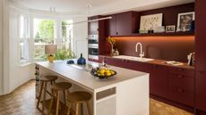 A maroon and white kitchen with a large island in the middle and three wooden bar stools beside it. In the kitchen is also a large shelf of open shelving with artwork on top of it. There is also a view of the kitchen sink as well as built in appliances.