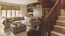 A cozy living room with exposed wooden staircase, large L-shaped sofa, patterned coffee table and brown armchair. Behind is a large wooden cabinet and built-in bookshelves. To the right is a bright window with a patterned statement blind seen.