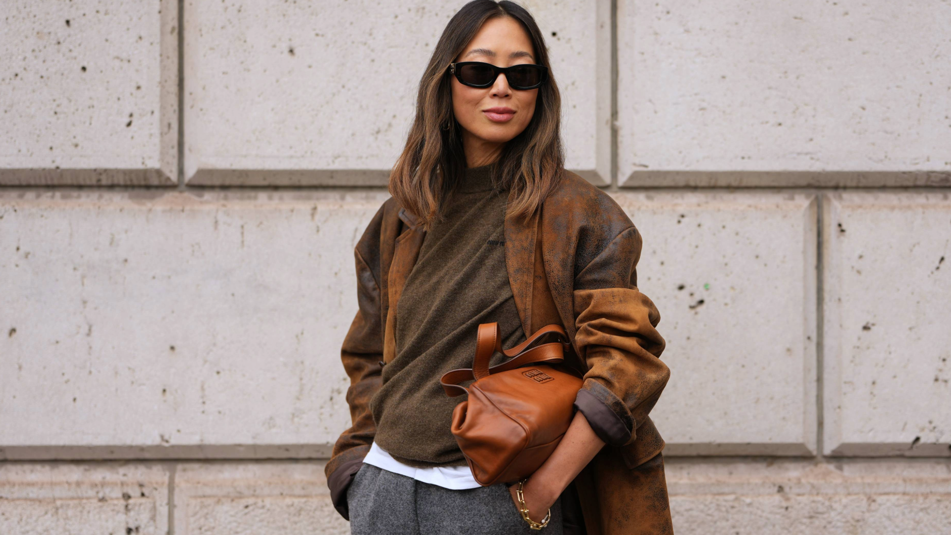 PARIS, FRANCE - OCTOBER 06: Aimee Song wears long wavy dark brown hair with center part, black rectangular sunglasses, small gold hoop earrings, a gold chain bracelet, a brown leather shoulder bag, a distressed brown suede oversized blazer jacket, an olive-green wool crewneck sweater, a white cotton T-shirt with visible hem, grey wool wide-leg trousers, outside Miu Miu, during Paris Fashion Week - Womenswear Spring Summer 2026, on October 06, 2025 in Paris, France (Photo by Edward Berthelot/Getty Images)
