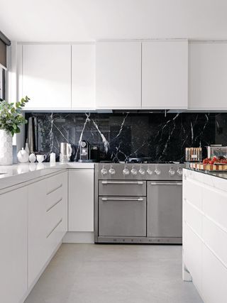 An all-white kitchen (white floors, white cabinets, white counter tops, and a white island) with a black marble backsplash and a large, silver oven