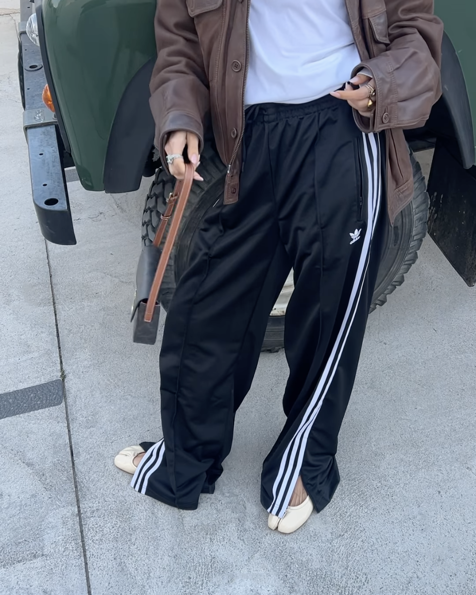 style influencer stands next to a car posing with a brown leather jacket, white tee, Adidas track pants, and Margiela ballet flats