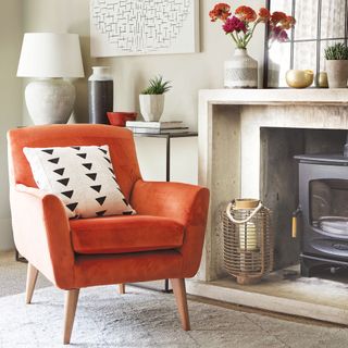 a neutral living room with a white mantle surrounding a black woodburner, with a bright orange armchair in front