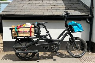 The Tern GSD S10 cargo bike fully loaded pointing to the right with a full cargo load. There is a blue bag on the front and a wooden crate on the back full of shopping. The bike is in front of a black and white building which has a very low tiled roof