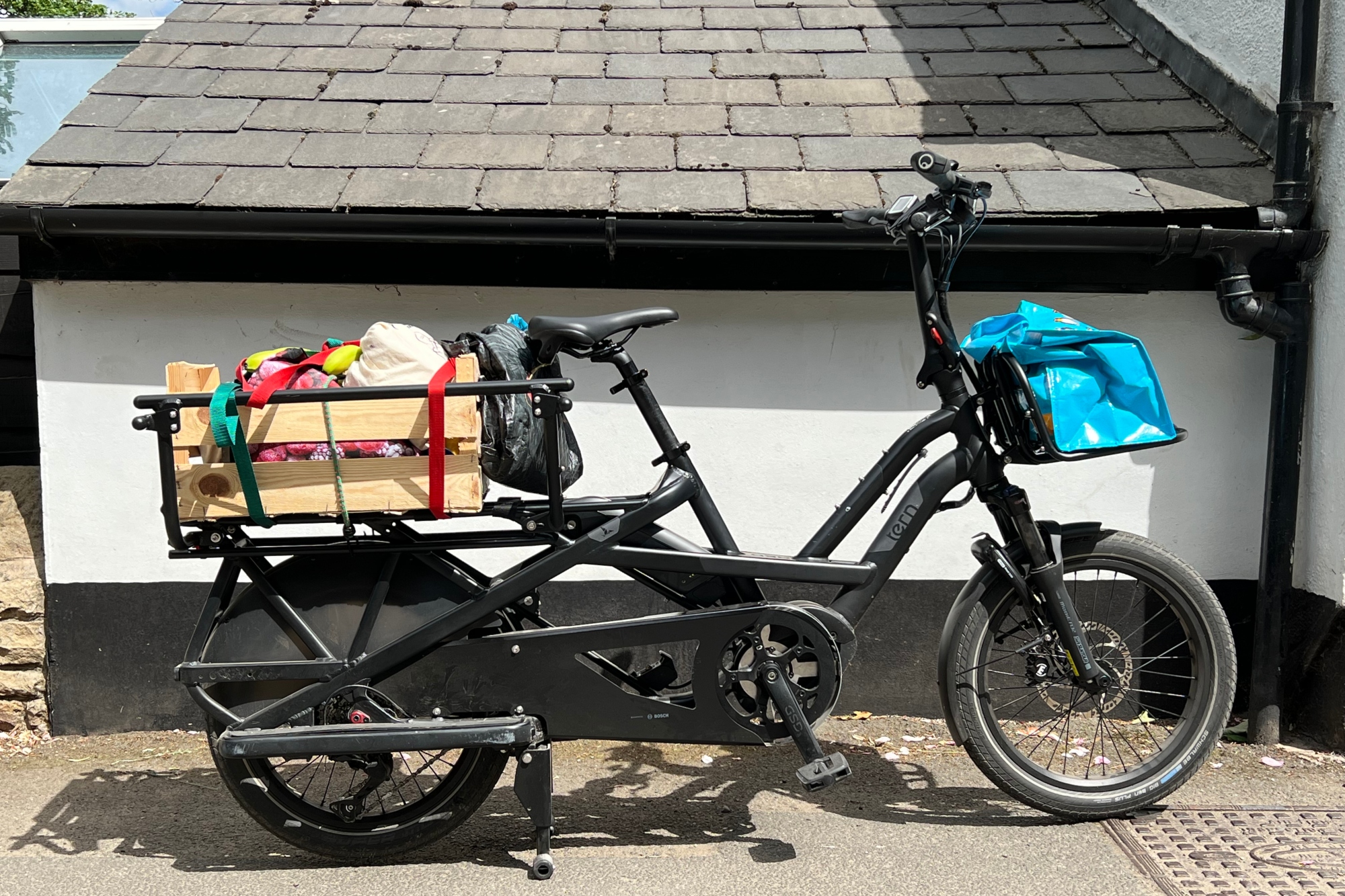 The Tern GSD S10 cargo bike fully pointing to the right with a full cargo load. There is a blue bag on the front and a wooden crate on the back full of shopping. The bike is in front of a black and white building which has a very low tiled roof