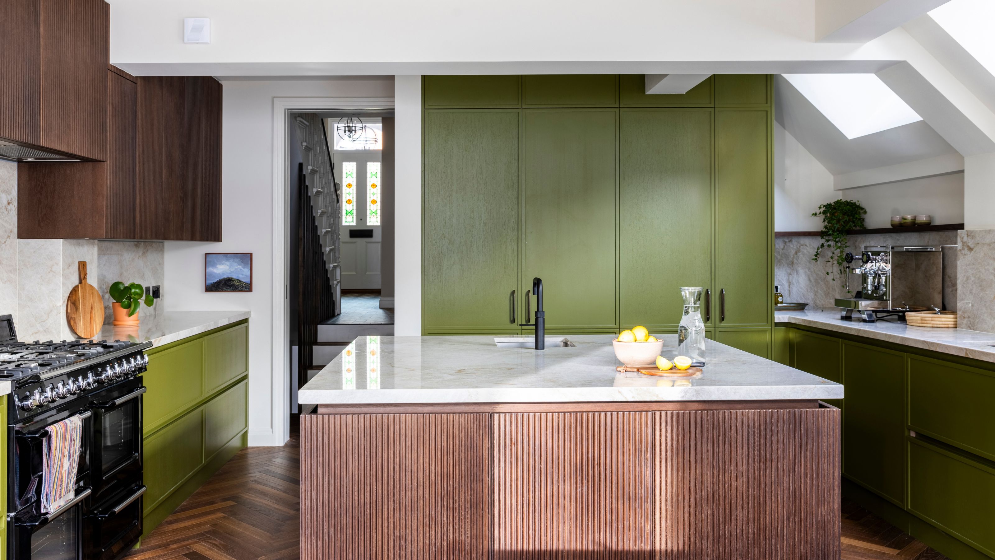 A modern kitchen with warm olive green cabinets, dark wood kitchen island and wall units, white walls, and parquet flooring.