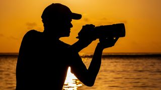 A silhouette of a person holding the LUMIX S1RII against a vibrant orange sunset over a tranquil ocean, capturing a serene moment