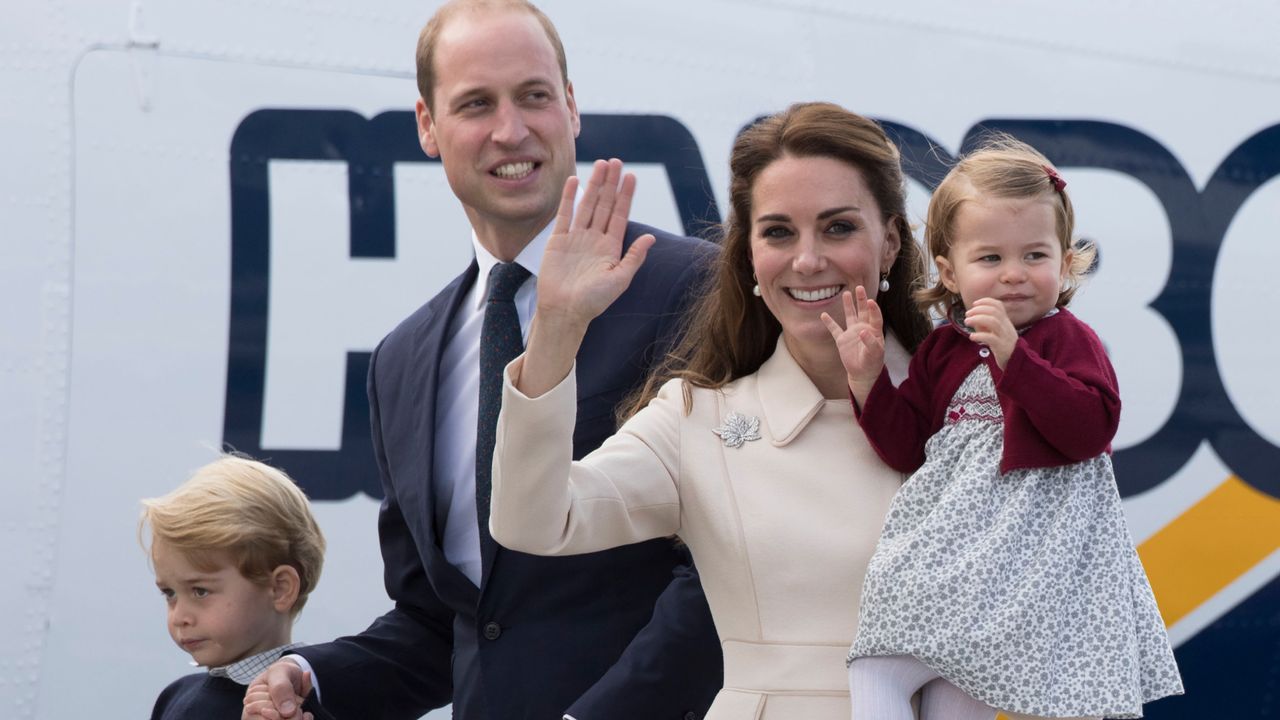 Kate Middleton waving and smiling holding a baby Princess Charlotte standing next to Prince William, wearing a blue suit and holding hands with toddler Prince George