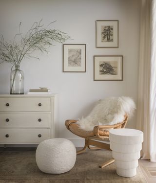 A corner of a bedroom with a wooden chair, white pouffe and side table, next to a cream chest of drawers