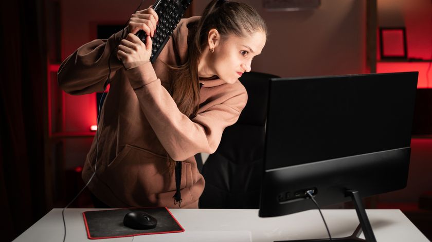 A woman standing by a desk with a desktop computer setup with a monitor and mouse. She is holding the keyboard as if ready to swing it at the monitor.