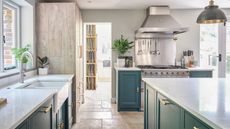 A light and airy kitchen with pendant lights, blue units and flagstone floor. A stainless steel backsplash and extractor fan behind/above the stove. 