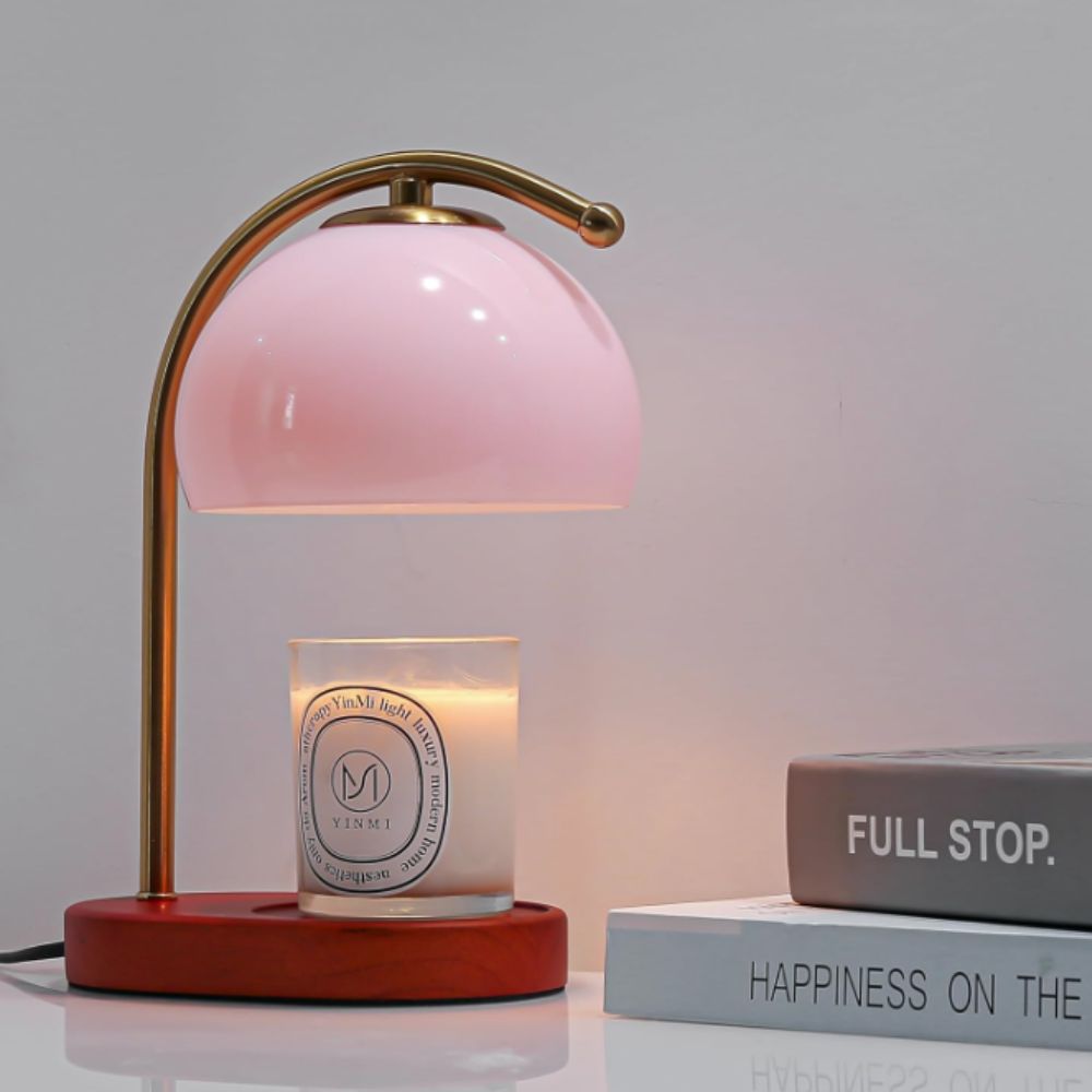 Retro candle warmer with pink glass shade and curved gold stem on a wooden base with a candle on