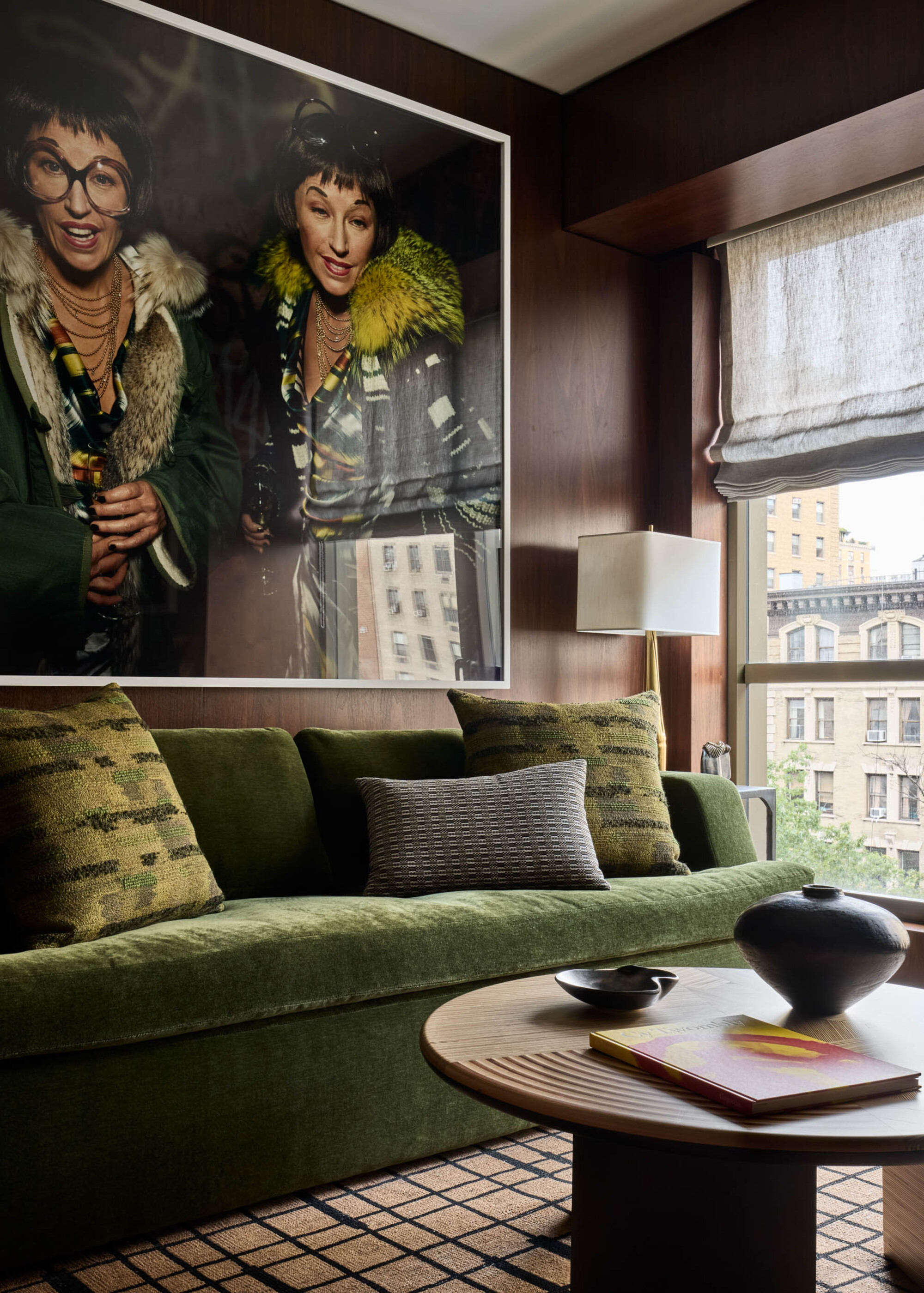 Image of a dark wood living room with a green velvet sofa that has a photography art piece with a similar color scheme above it. The window has sheer, gray Roman blinds.