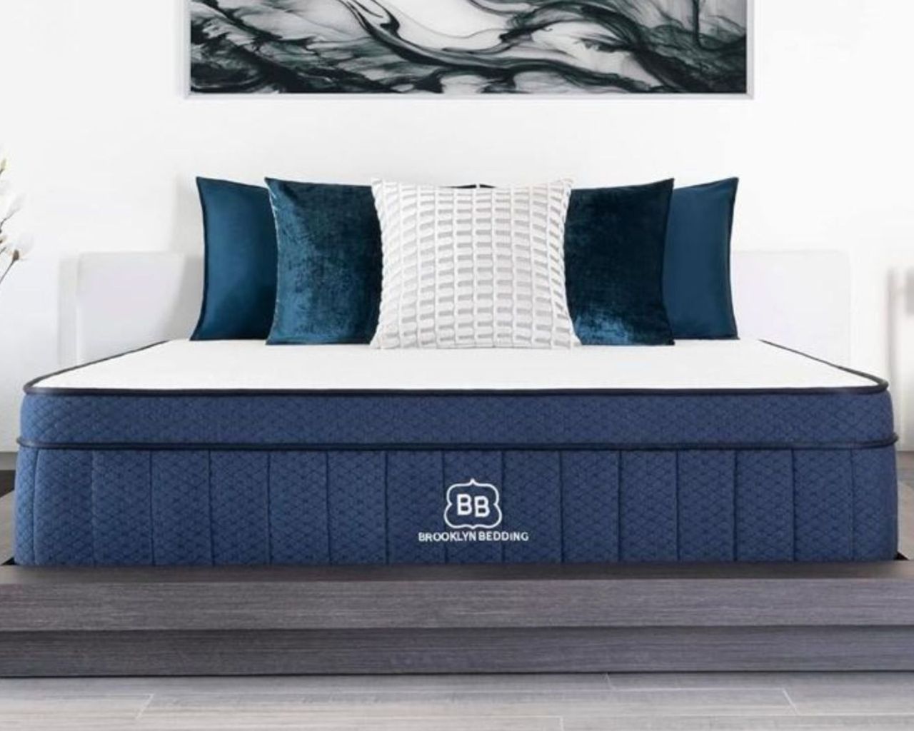 A blue Brooklyn Bedding Aurora Luxe mattress with white and blue cushions with white walls and a large artwork above, and light wooden flooring.