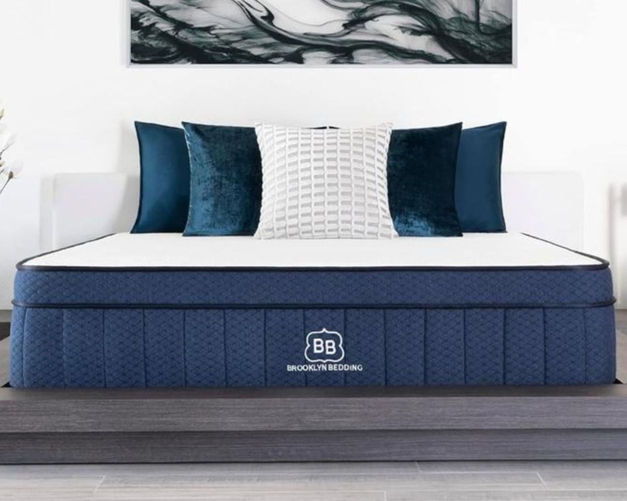 A blue Brooklyn Bedding Aurora Luxe mattress with white and blue cushions with white walls and a large artwork above, and light wooden flooring.