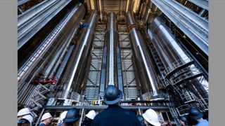 photo looking up at four huge stainless-steel cylinders inside an open-air high bay, with a crowd of people wearing hard hats in the foreground.