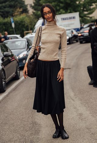 Milan Fashion Week spring 2025 street style photo of a woman wearing geek chic trend in the form of a tan turtleneck, black pleated skirt, black stockings and black tabi toe shoes