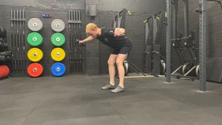 Harry Bullmore performs a rear delt fly with dumbbells in a gym. He is hinging forward at the hips so that his torso is angled about 45 degrees upwards from the floor. He holds light dumbbells in his hands and maintains a slight bend in his elbow as he holds them out to either side. Behind him we see weights plates, air bikes and other gym equipment.