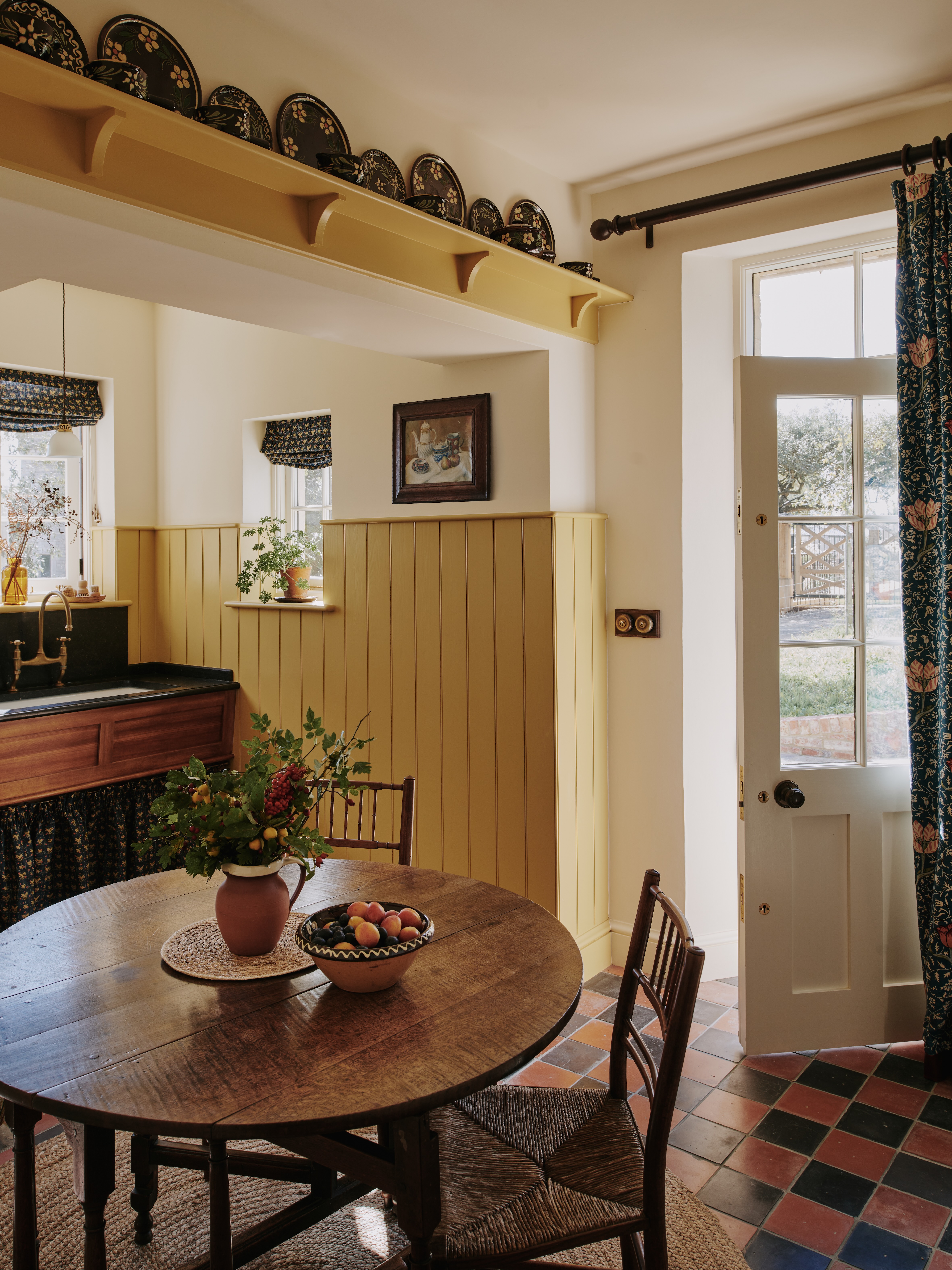 Corner of a kitchen with red and black square tiles, yellow panelling and a dark brown circular table and chairs