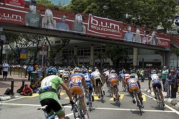 The peloton pass underneath a B.U.M Equipment banner in Kuala Lumpur. B.U.M Equipment is a Malaysian clothes range.
