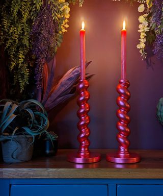 Two pink twisted tall candlesticks, on a wooden table with plants in the background. Ambiance is dark and moody