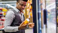 photo of a man checking the label on a yellow can that he's pullled from a fridge at a large grocery store