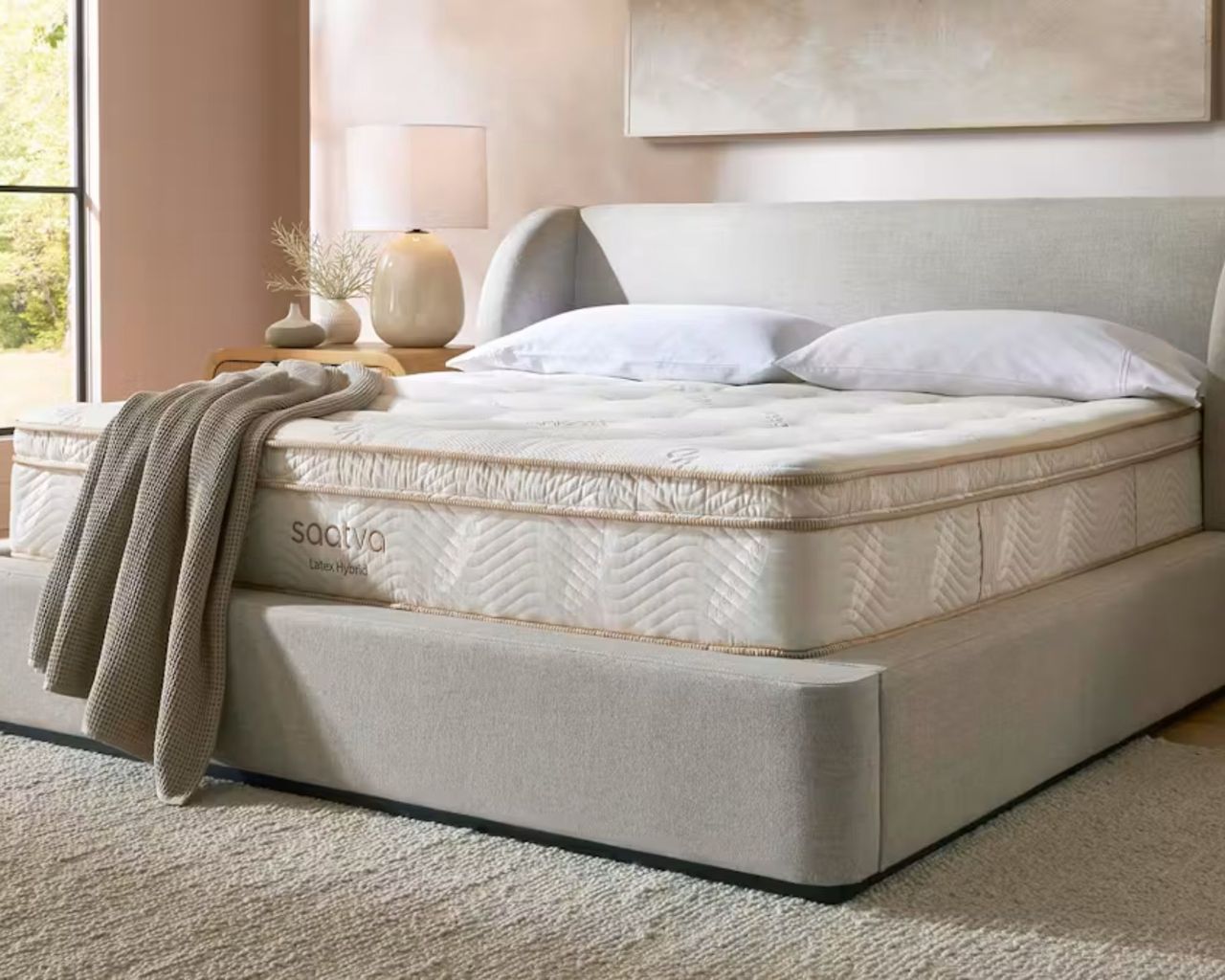 A white Saatva Latex Hybrid Mattress on a grey bed in a blush pink bedroom with a large cream rug, large artwork, and large windows.