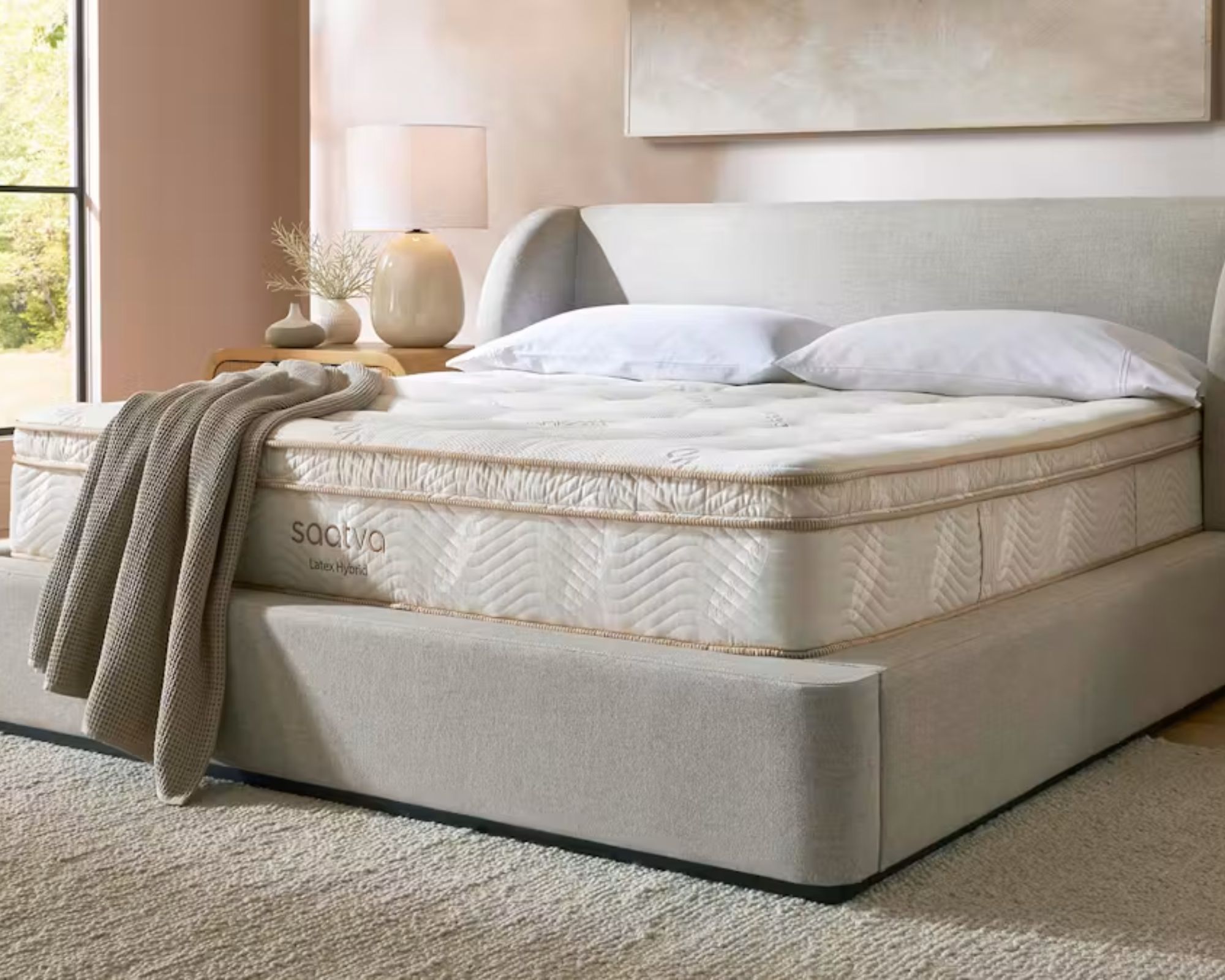 A white Saatva Latex Hybrid Mattress on a grey bed in a blush pink bedroom with a large cream rug, large artwork, and large windows.