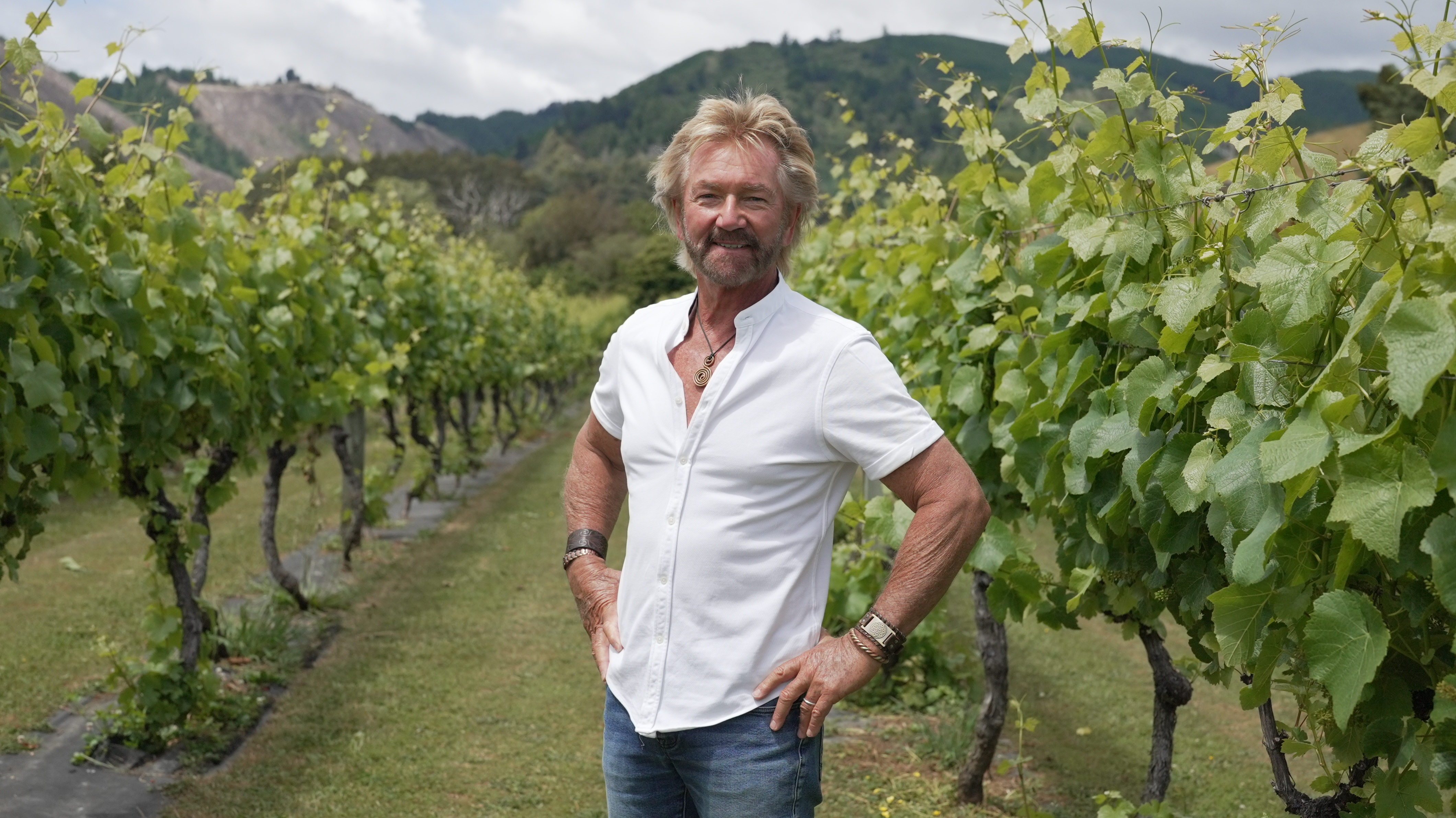 Noel Edmonds smiling with a short-sleeved white shirt on and standing in his New Zealand vineyard. 