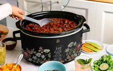 A black slow cooker on a kitchen island with a wildflower pattern, surrounded by ingredients 