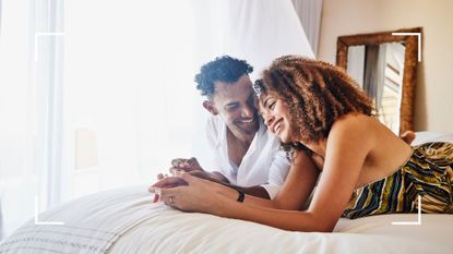 Woman and man representing flatiron sex position, lying down on white bedsheets laughing and smiling together