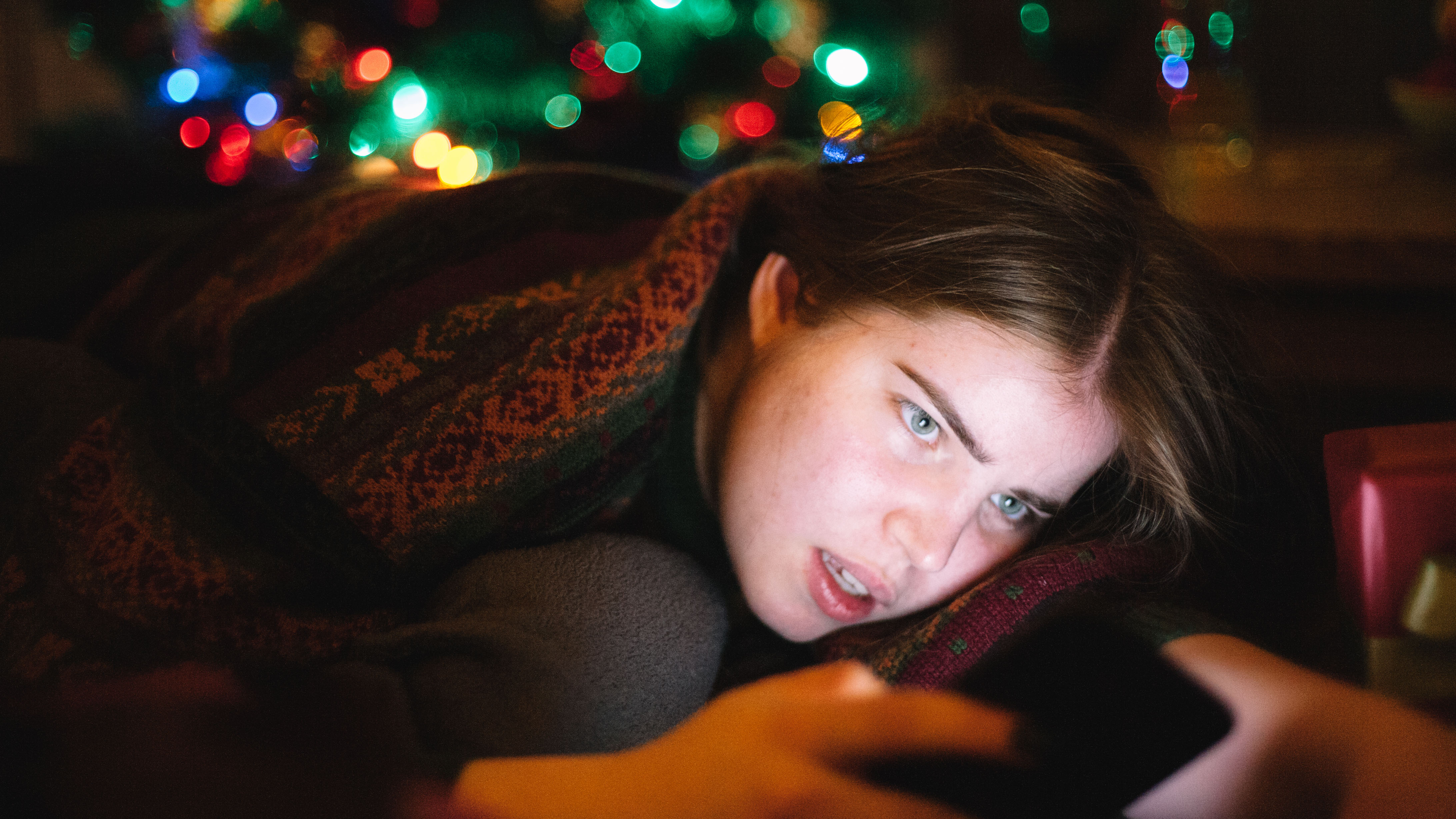 Young woman using smart phone while lying on the sofa at home with glowing Christmas tree on the background
