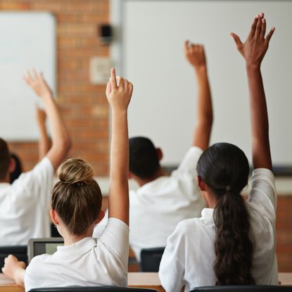 Young girls in a classroom, raising their hands. 