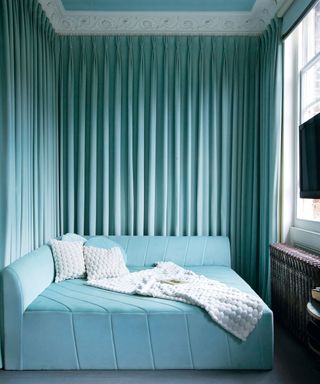 Guest room with light turquoise velvet square sofa bed with diagonal seams, and pleated curtain around the walls