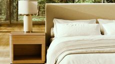 Saatva bed with white bedding beside a wooden nightstand with a white lamp on top. The flooring is light wood, and behind is a forest landscape seen through large windows.