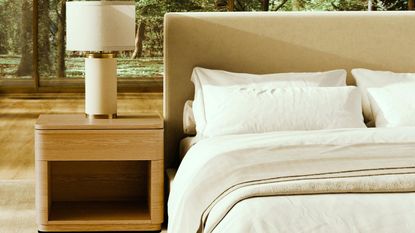 Saatva bed with white bedding beside a wooden nightstand with a white lamp on top. The flooring is light wood, and behind is a forest landscape seen through large windows.