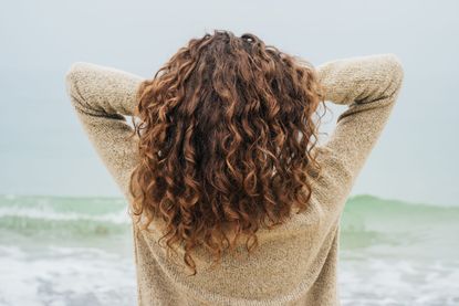 Curly brunette in a sweater on the coast faces the sea and straightens hair, view from the back