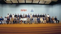 a large group of people in suits sits on a stage with orange carpet