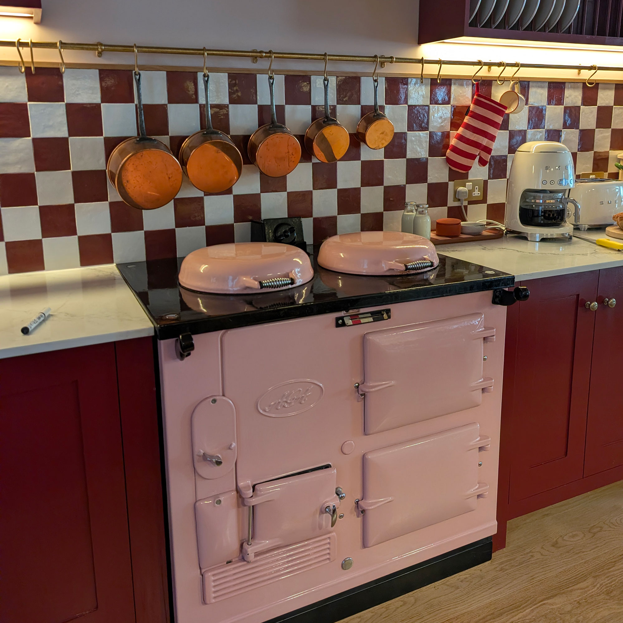 Pink AGA in kitchen with red cabinetry beneath white and red chequered tile splashback