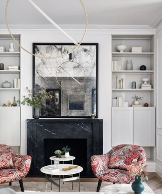 Small living room lighting ideas with pink floral armchairs, white walls, black framed tarnished mirror and brass hoop light fitting with LED bar across center