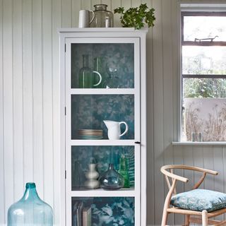 Tall cabinet with wallpapered interior in front of white panelled wall