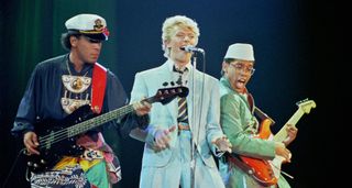 David Bowie wears a powder blue suit as he is flanked onstage by Carlos Alomar with a red Stratocaster and Carmine Rojas on bass guitar. Rojas ears a captain's hat.