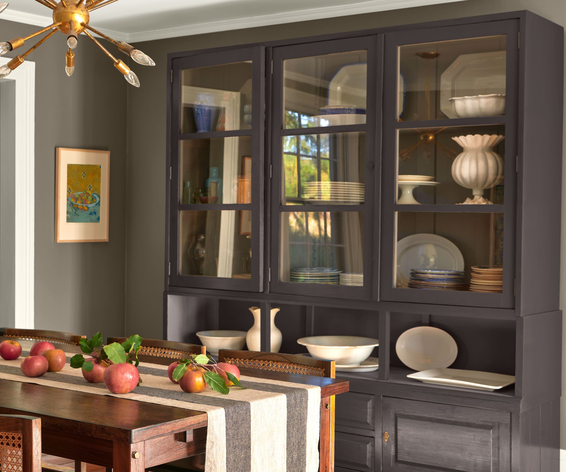 Dining room with a large dresser painted in black bean soup by Benjamin Moore, a wooden dining table with a striped runner and pomegranates on top, and grey walls.